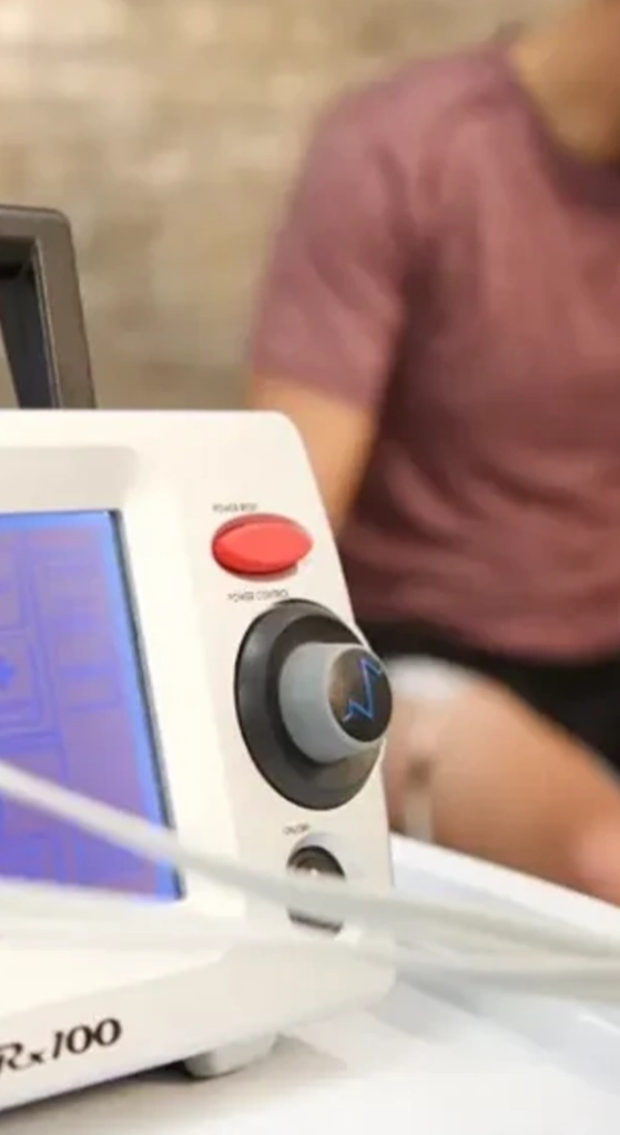 ARP neuro therapy device in the foreground while a patient receives electrical stimulation treatment in a rehab setting.